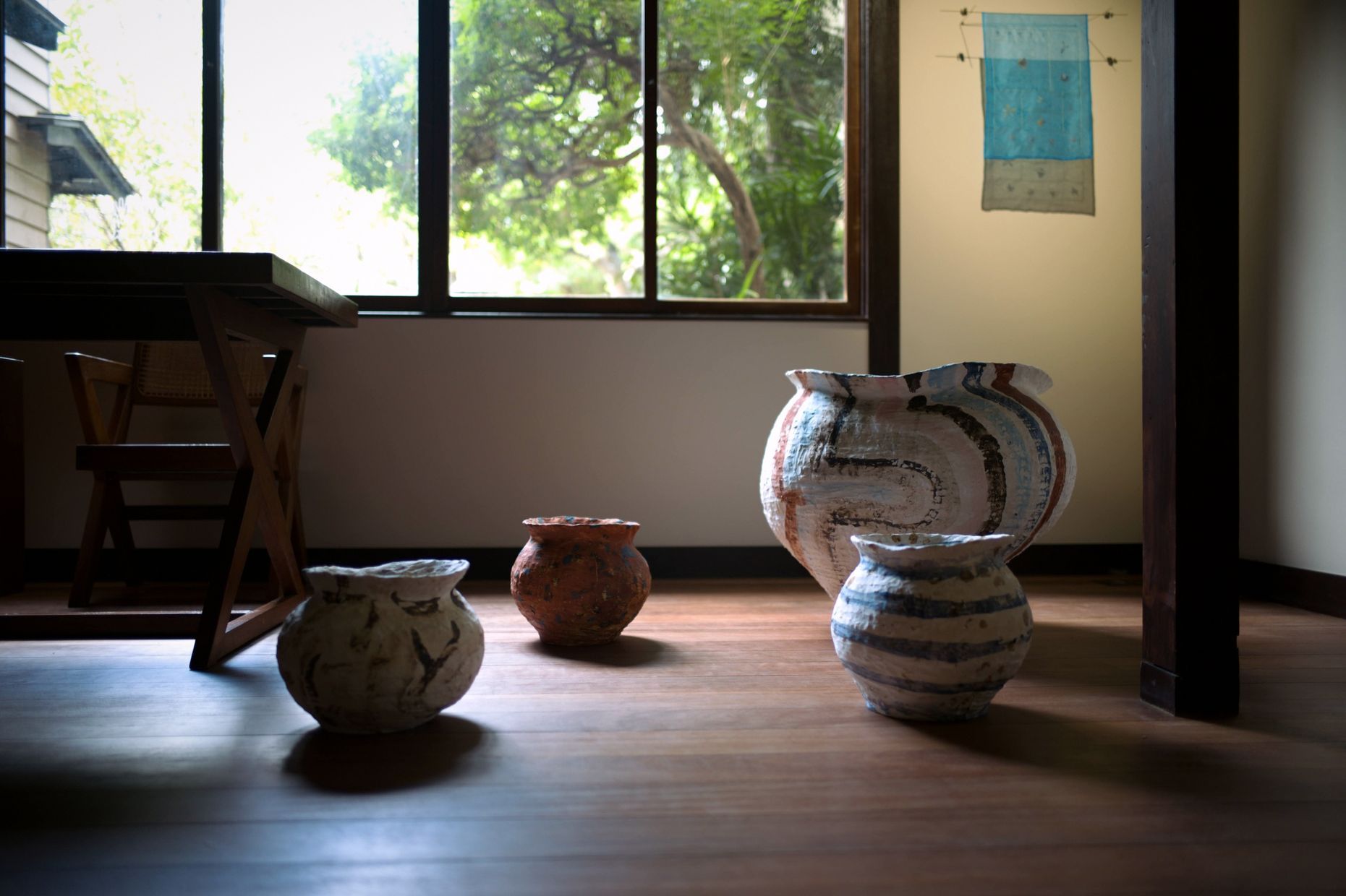 Four ceramic jars on a sunlit wooden floor before a window onto trees, with a blue textile on the wall.