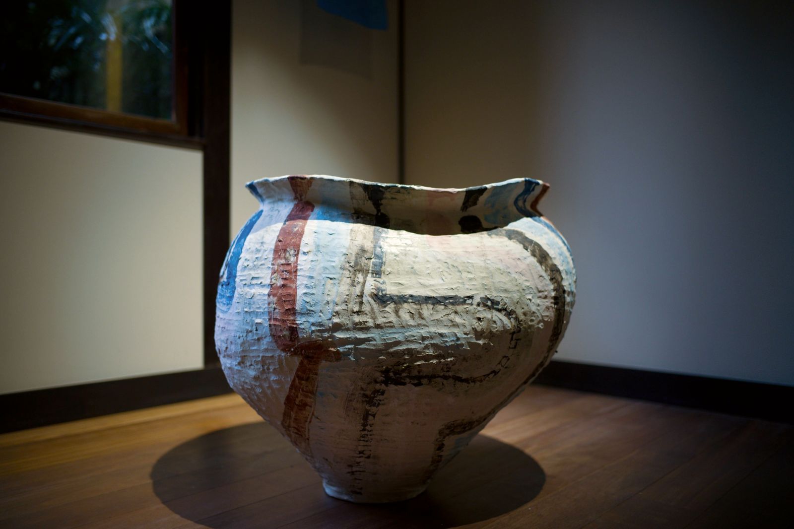 A large hand-built jar with red, blue and black painted lines on a wooden floor.