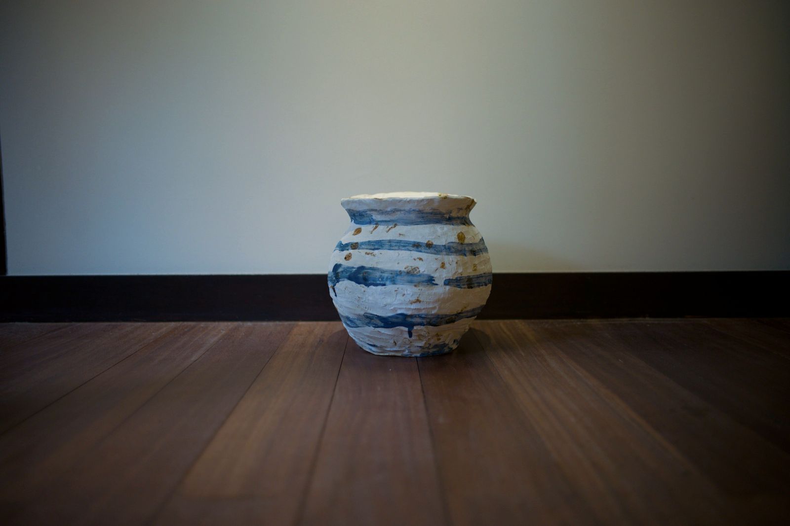 A white jar with blue bands on a dark wooden floor against a pale wall.