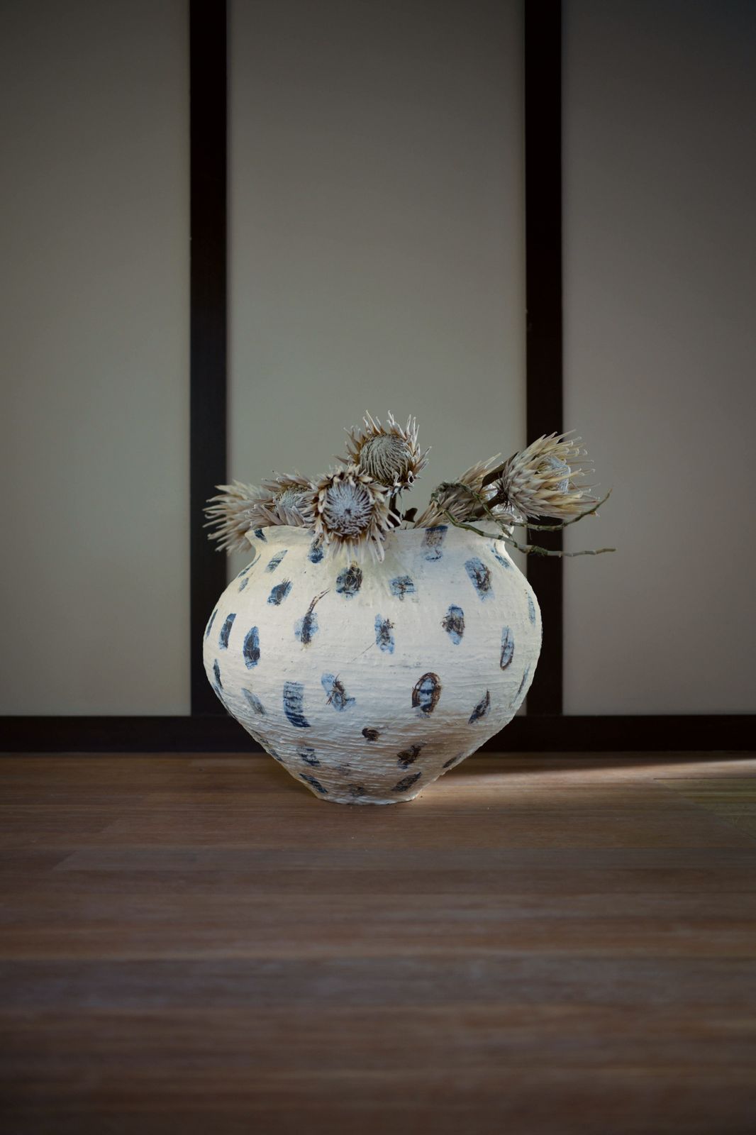 Dried protea flowers in the speckled jar on a wooden floor before dark wooden posts.