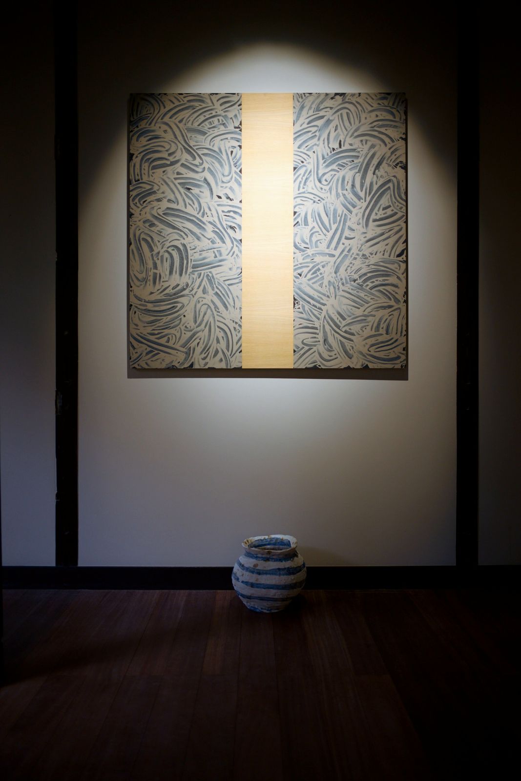 A square work of swirling blue mud marks with a vertical band of bare wood, lit by a spotlight, with a striped jar on the floor below.