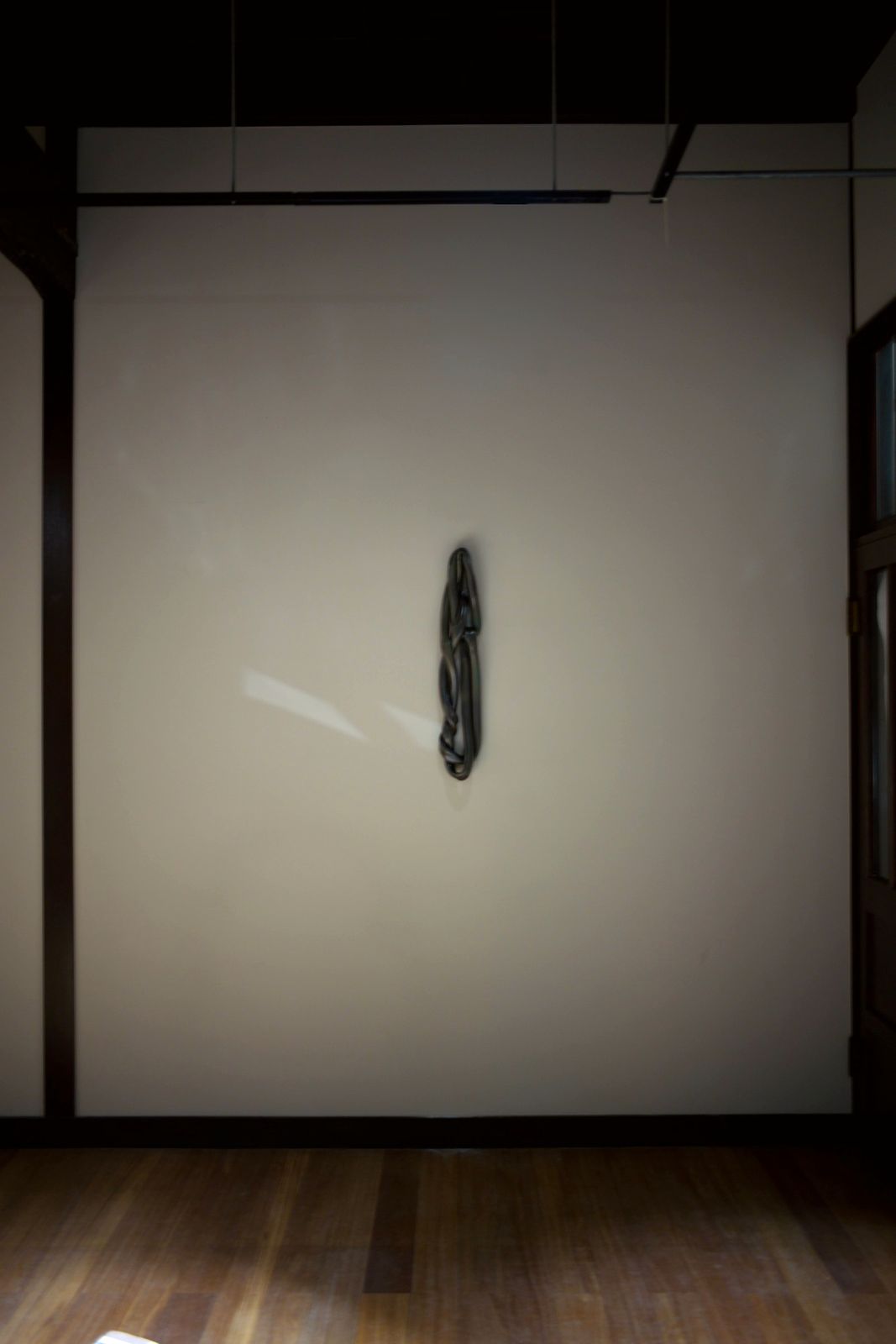 A tall, dark knotted ceramic form hanging on a pale wall under a spotlight.