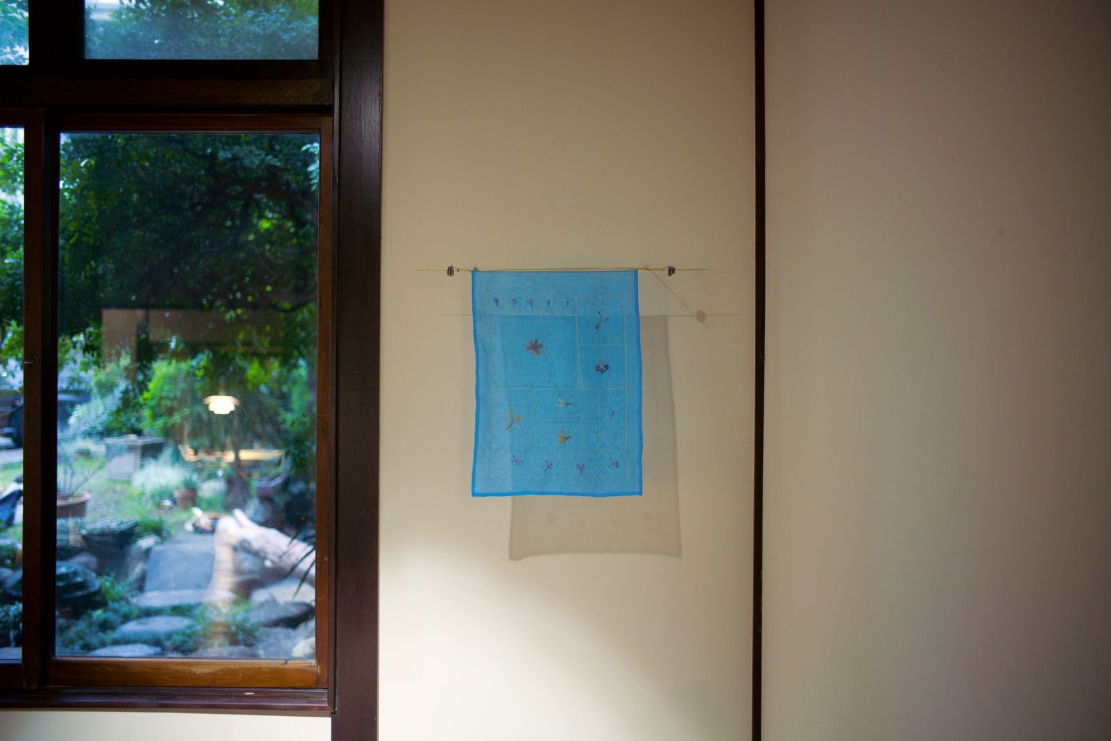 A sheer blue textile hanging on a wall beside a window onto a garden.