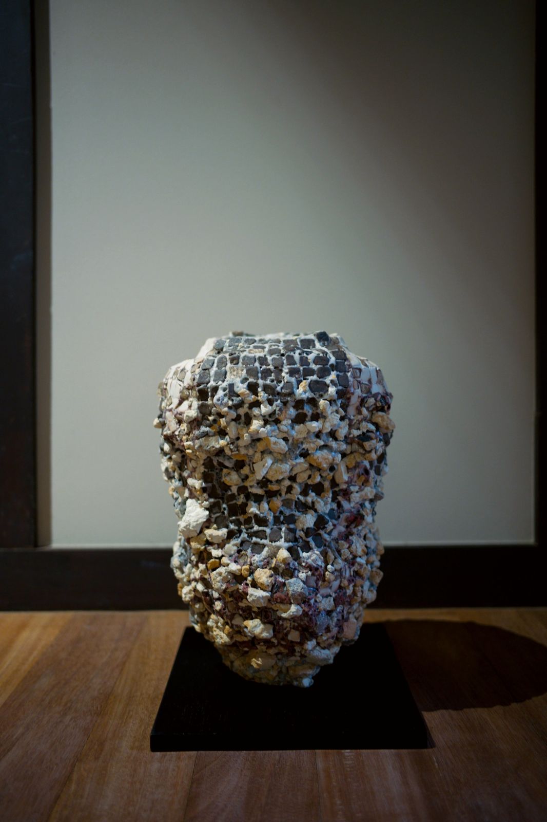 A tall vessel covered in mosaic-like fragments on a black plinth against a pale wall.